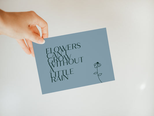 hand holding a blue greeting card that says "flowers can't grow without a little rain" with a sketched flower next to the words