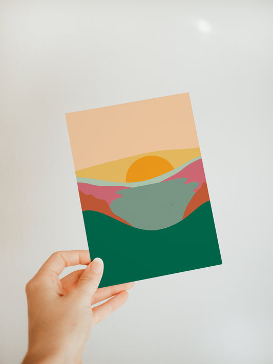 hand holding a greeting card with a blocky landscape with the sun rising over it design 