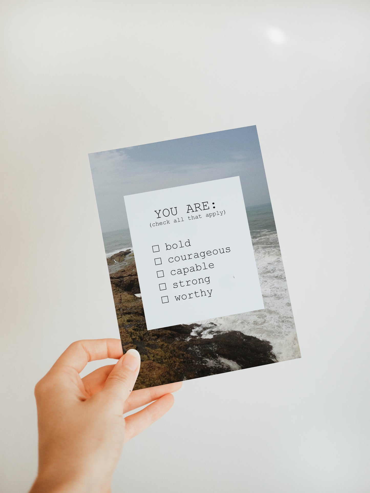 hand holding a greeting card with an ocean scene in the background and a white square in the foreground that says "you are: (check all that apply) bold, courageous, capable, strong, worthy."