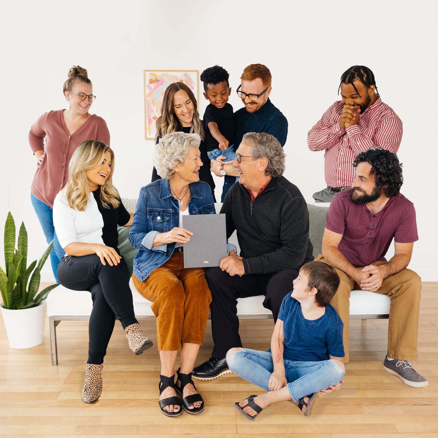 Group of people sitting on a wooden floor in a room with a plant and a white wall. Center couple is holding an IMPACT book and looking joyfully at each other. People surrounding them are smiling and looking excited to have given them this gift.