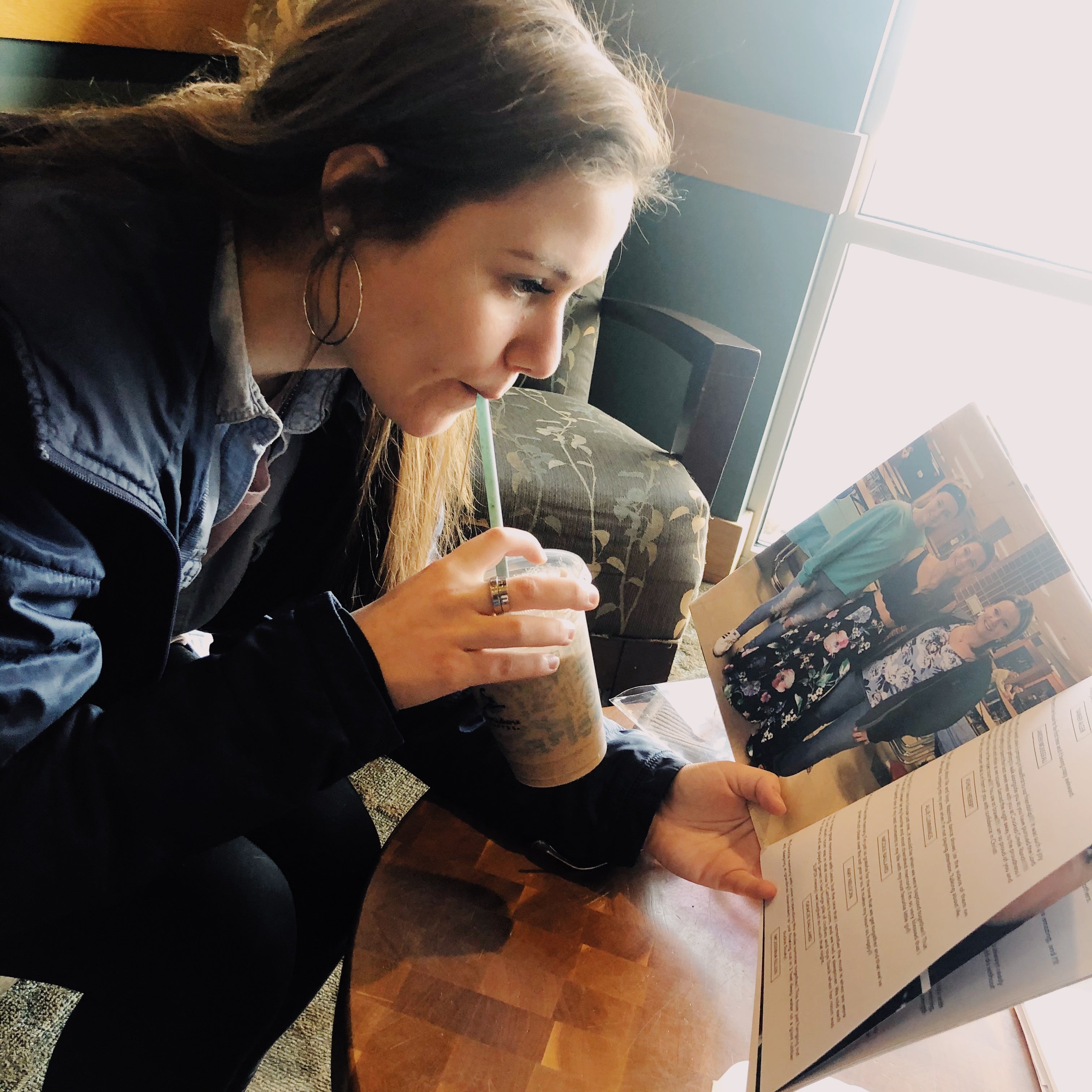 Woman sitting at a table reading a book with a drink in her hand.
