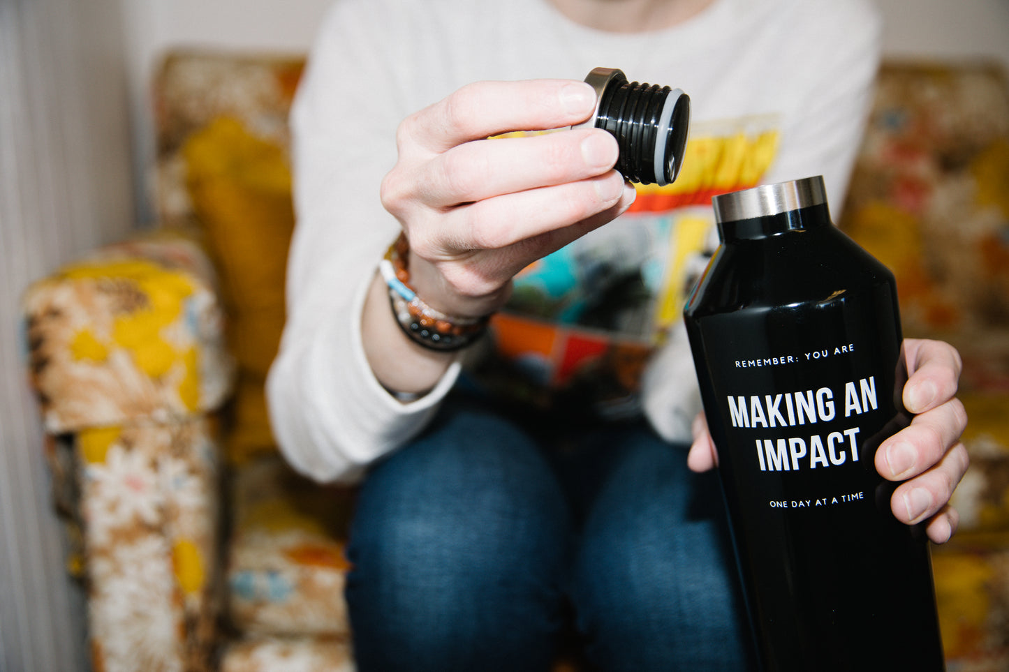 Person sitting on a couch holding a black stainless steel water bottle in one hand and the screw off lid in the other. The water bottle says "Remember, you are making an impact, one day at a time."