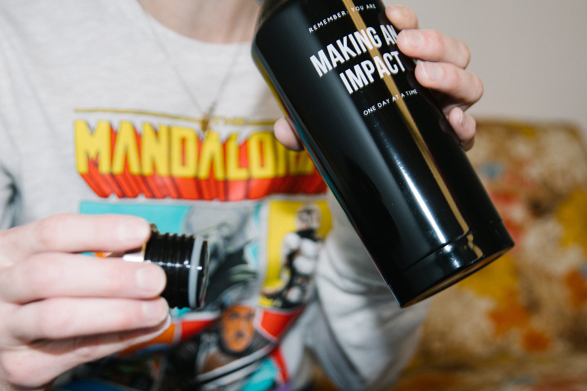 Person holding a black stainless steel water bottle in one hand and the screw off lid in the other lifting it toward the top of the frame to their mouth to take a drink. The water bottle says "Remember: you are making an impact one day at a time."