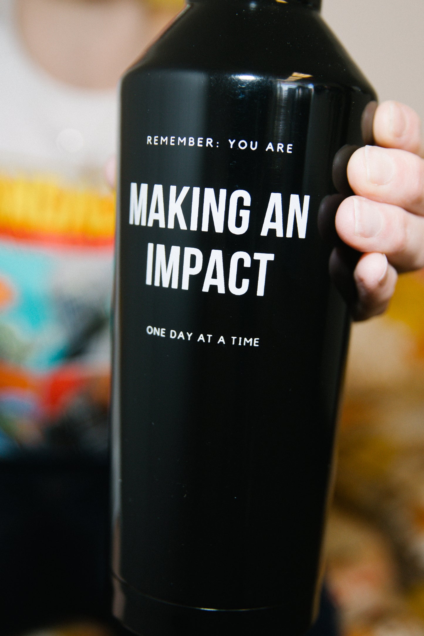 Person holding a black stainless steel water bottle out toward the camera in one hand. The water bottle says "Remember, you are making an impact, one day at a time."