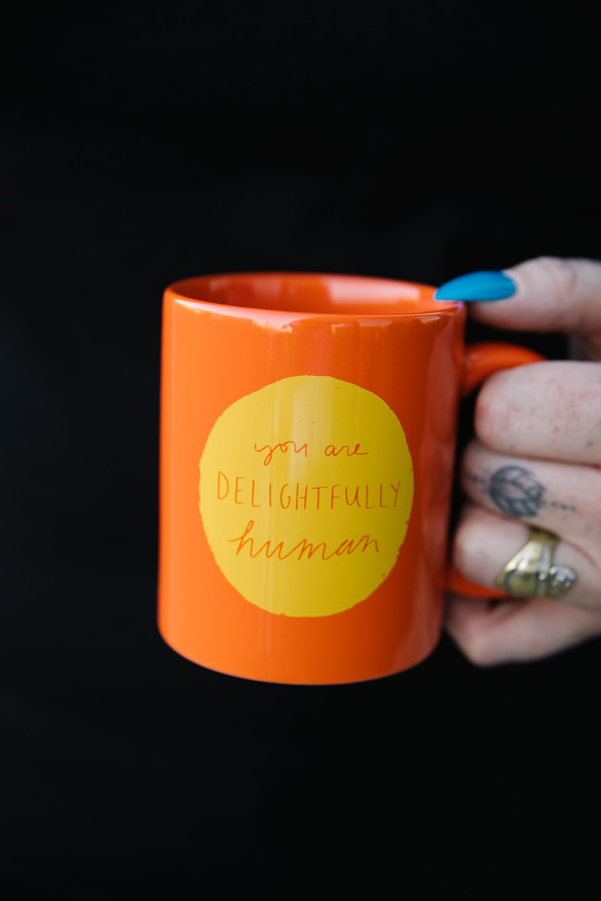 Hands holding a bright orange mug with a yellow circle in the center of the mug that says "you are delightfully human"