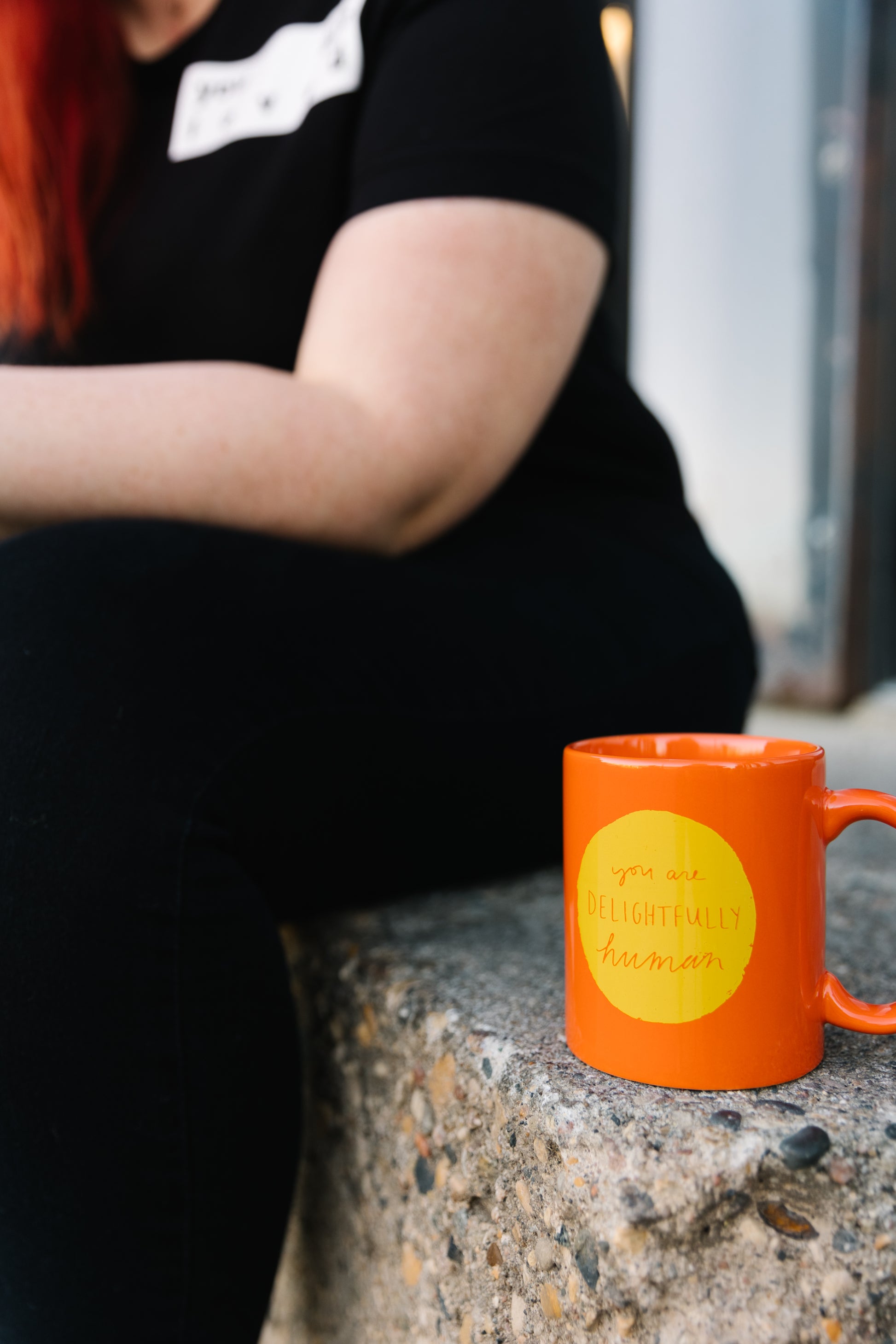 Person sitting on a step beside a bright orange mug with a yellow circle in the center of the mug that says "you are delightfully human"