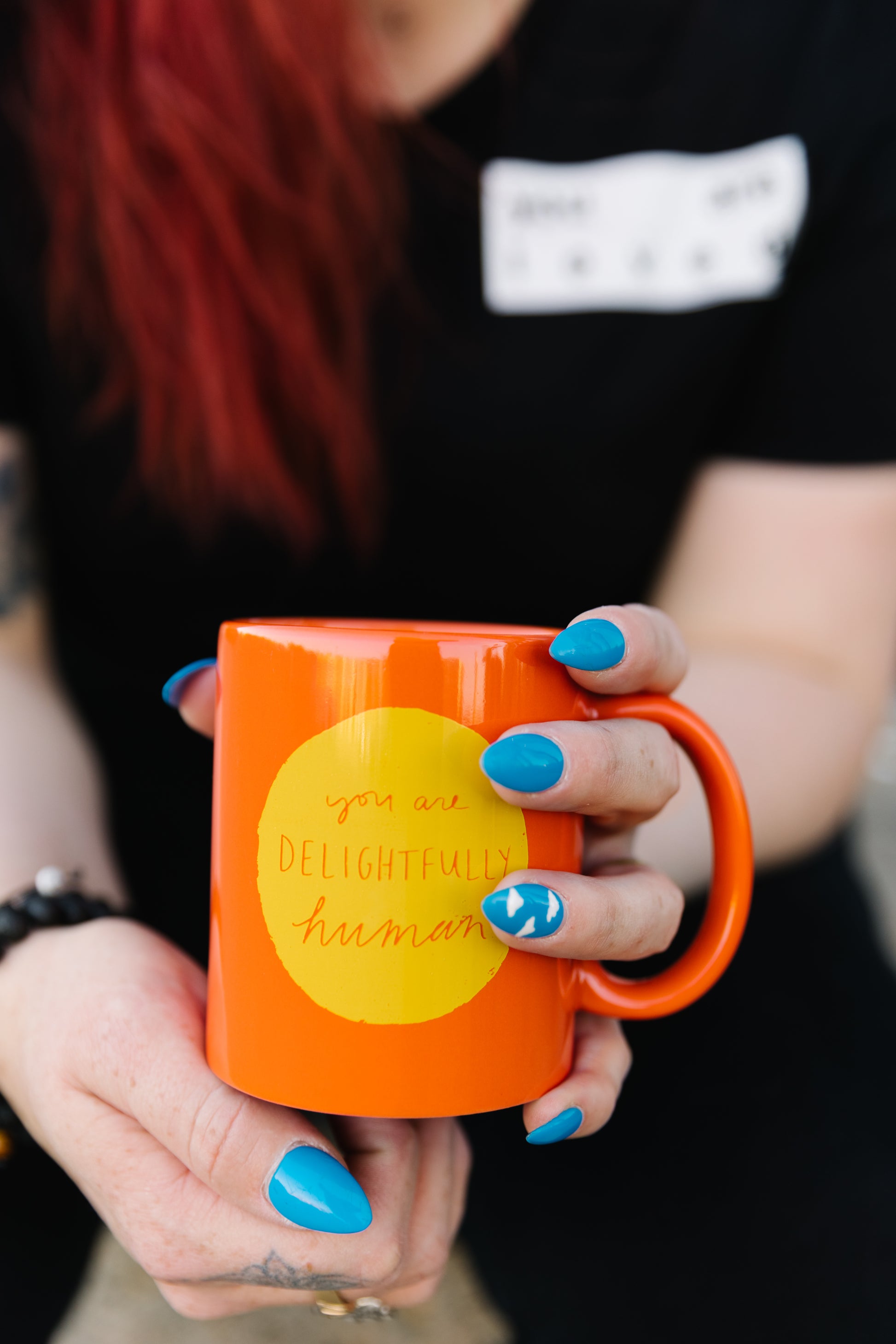 Hands holding a bright orange mug with a yellow circle in the center of the mug that says "you are delightfully human"