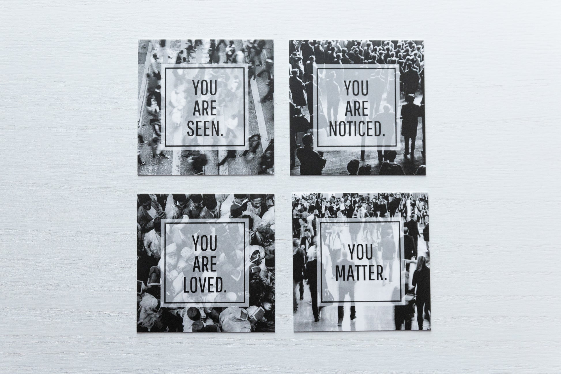 a set of four small square cards in black and white with a different view of a crowd of people on each card. Each set comes with one card with each of these sayings "you are seen", "you are noticed", "you are loved", "you matter". The back is left largely blank with only our website and hashtag in small font at the bottom, leaving plenty of room for a short note to be penned.