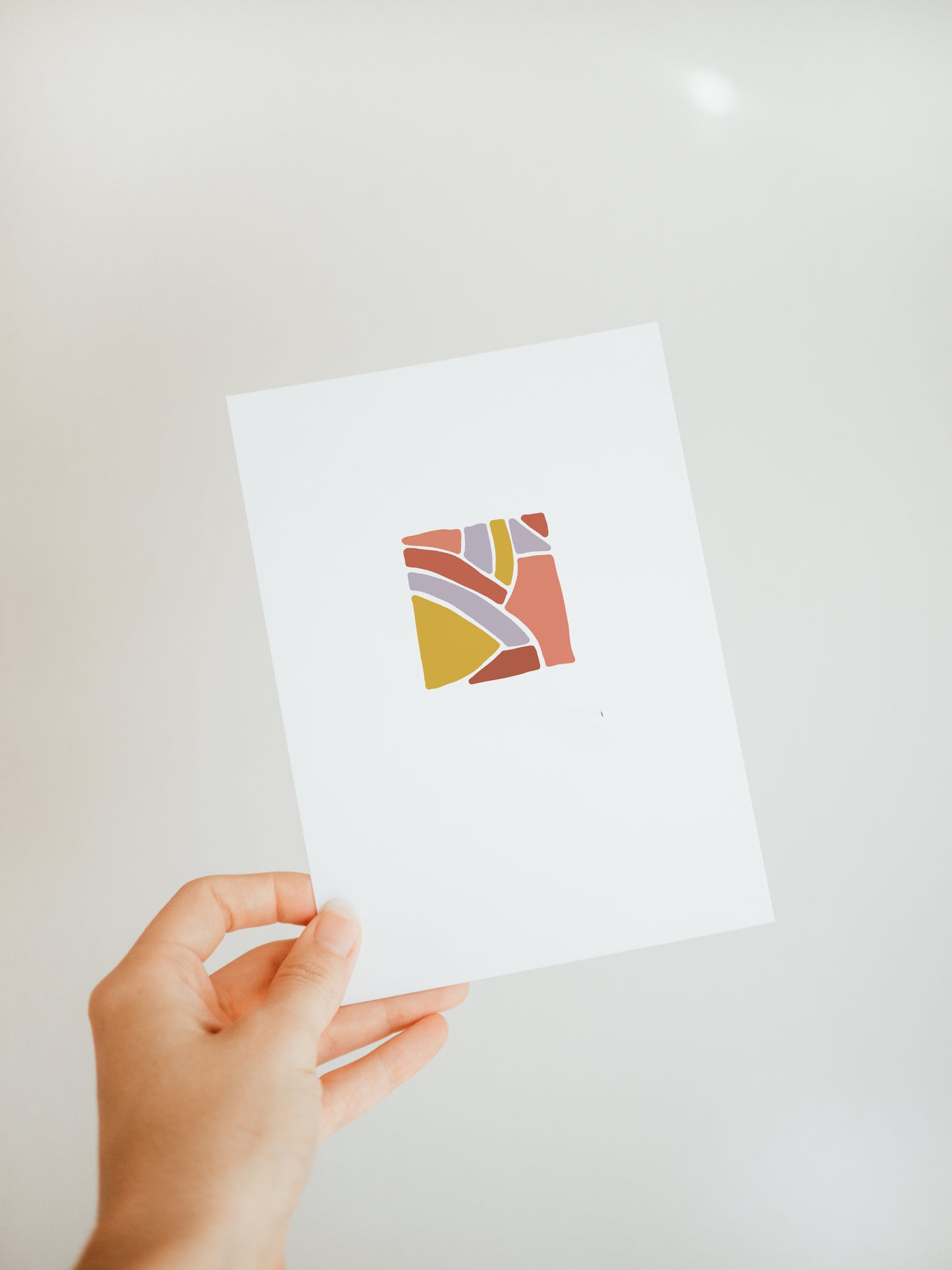 hand holding a white greeting card with a square design in the center made up of abstract shapes in muted yellow, salmon, brick, and lavender tones