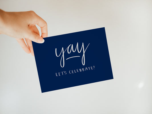hand holding a navy blue greeting card with white words saying "yay, let's celebrate!"