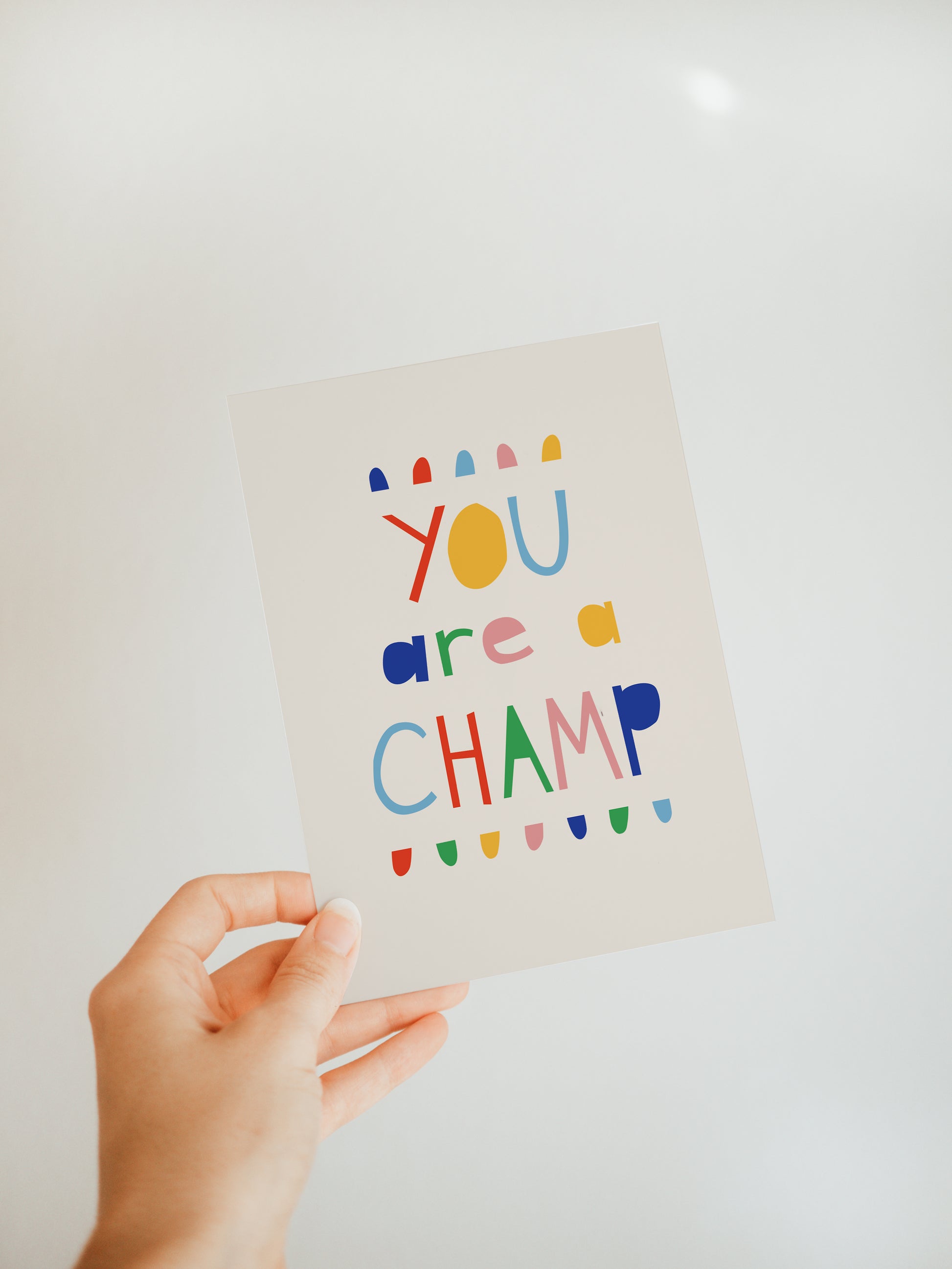 hand holding an off-white greeting card with a colorful banner along the top and bottom of words in colorful font that say "you are a champ"