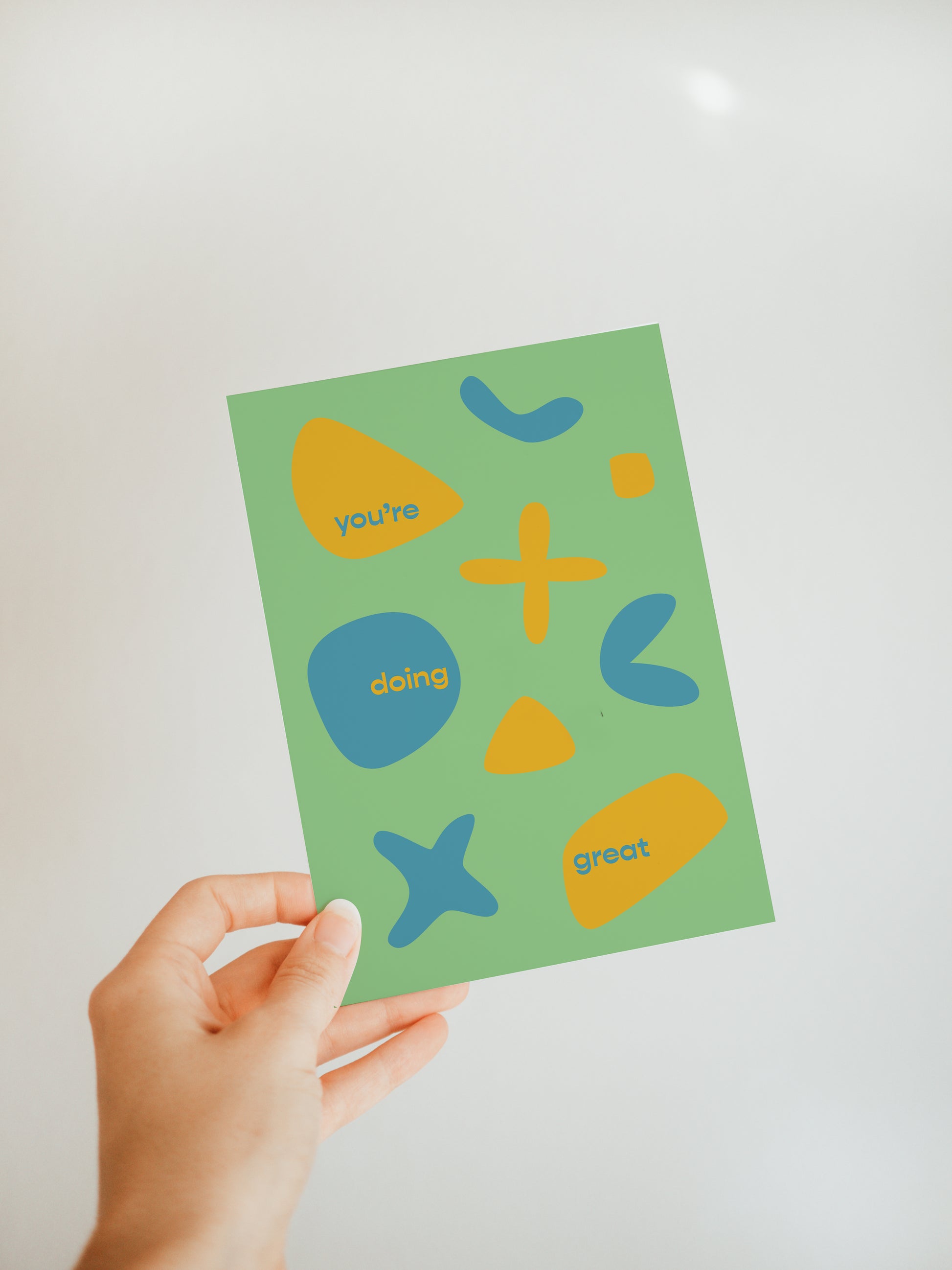 hand holding a green greeting card with the words "you're doing great" and abstract designs in yellow and blue