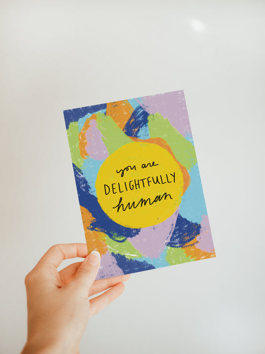 hand holding an abstract multicolored greeting card with the words "you are delightfully human" in the center in a yellow circle