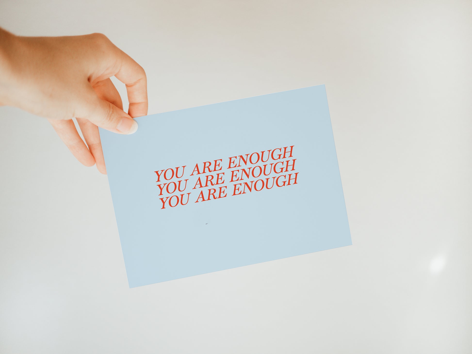 hand holding a light blue greeting card with the words "you are enough" repeated three times on the front in red font