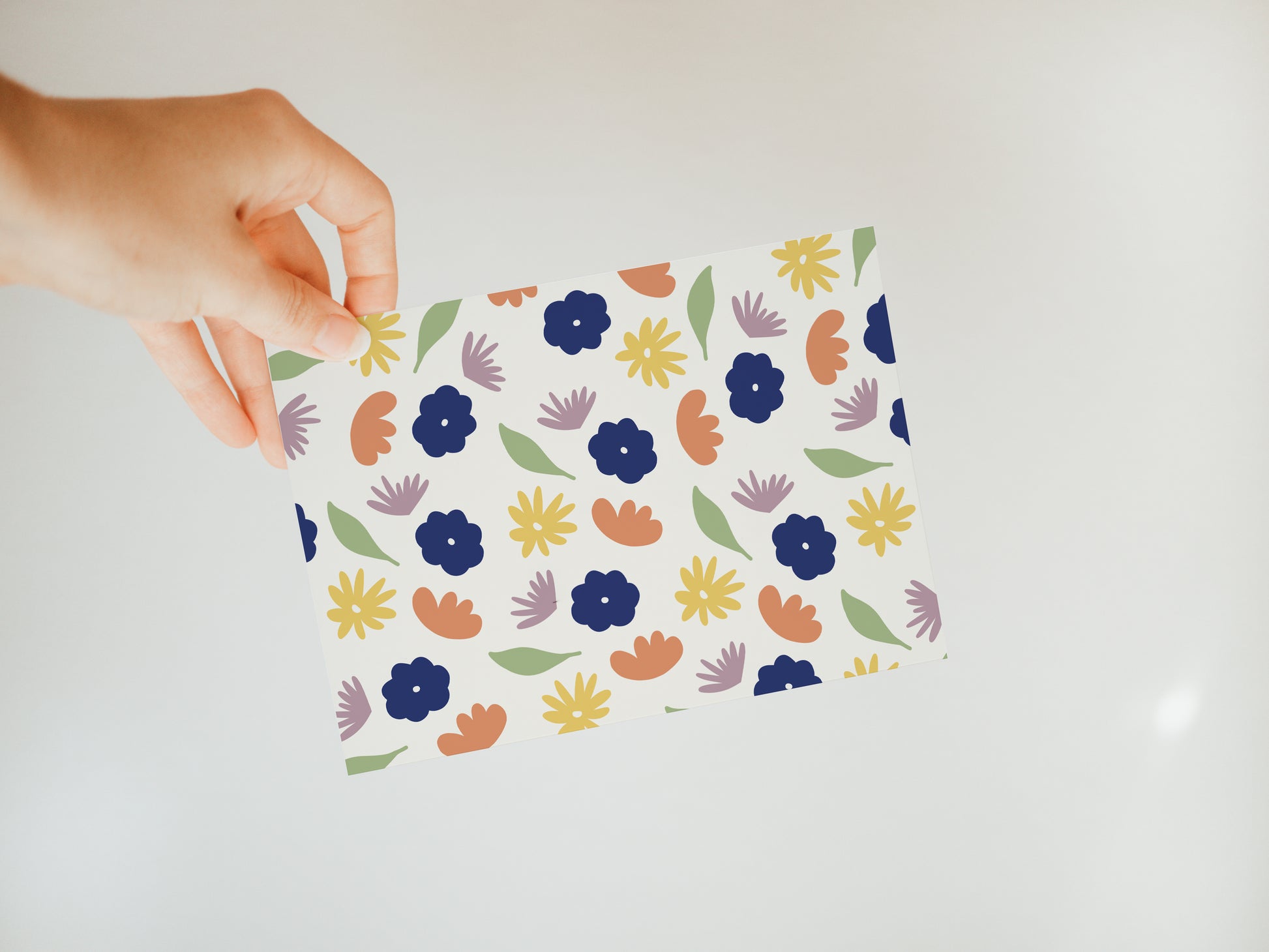 hand holding an off-white greeting card with a colorful floral and leaf pattern in green, dark blue, orange, yellow, and lavender.