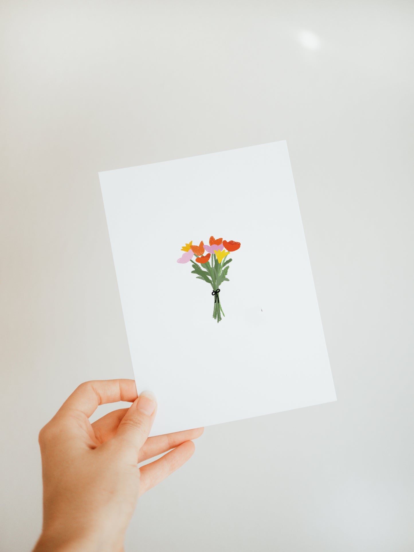 hand holding a white greeting card with a small and simple bouquet of yellow, pink, and red flowers with green stems tied with a black ribbon.