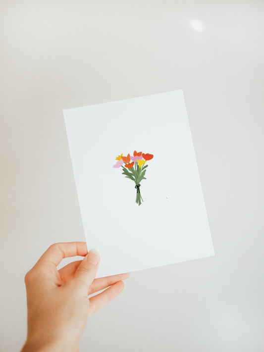 hand holding a white greeting card with a small and simple bouquet of yellow, pink, and red flowers with green stems tied with a black ribbon.