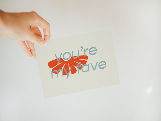 hand holding an off-white greeting card that says "you're my fave" in blue font with half of a flower in the background with the "o" as the flower's center