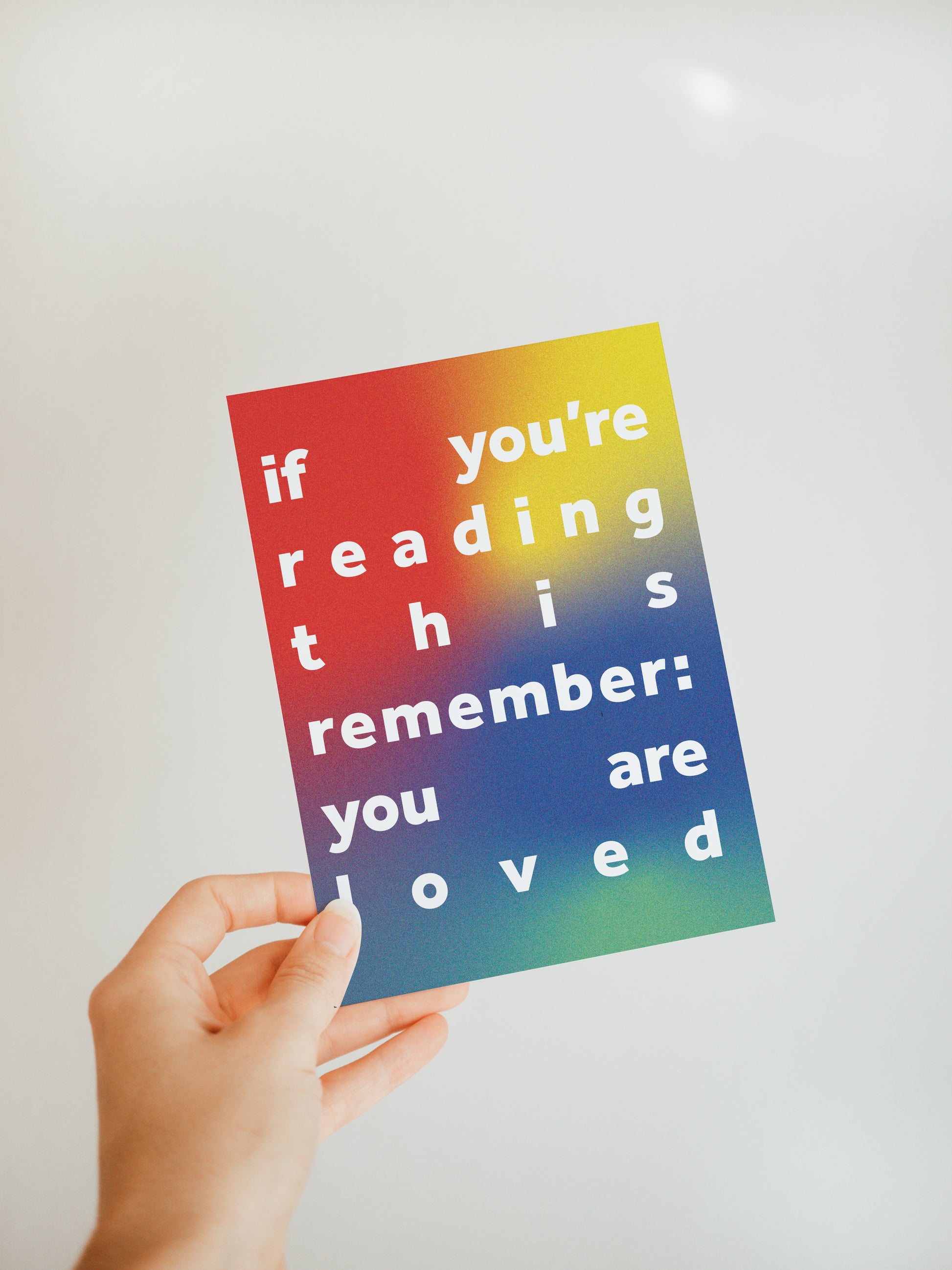 hand holding a multicolored greeting card that says "if you're reading this remember: you are loved" in white block letters