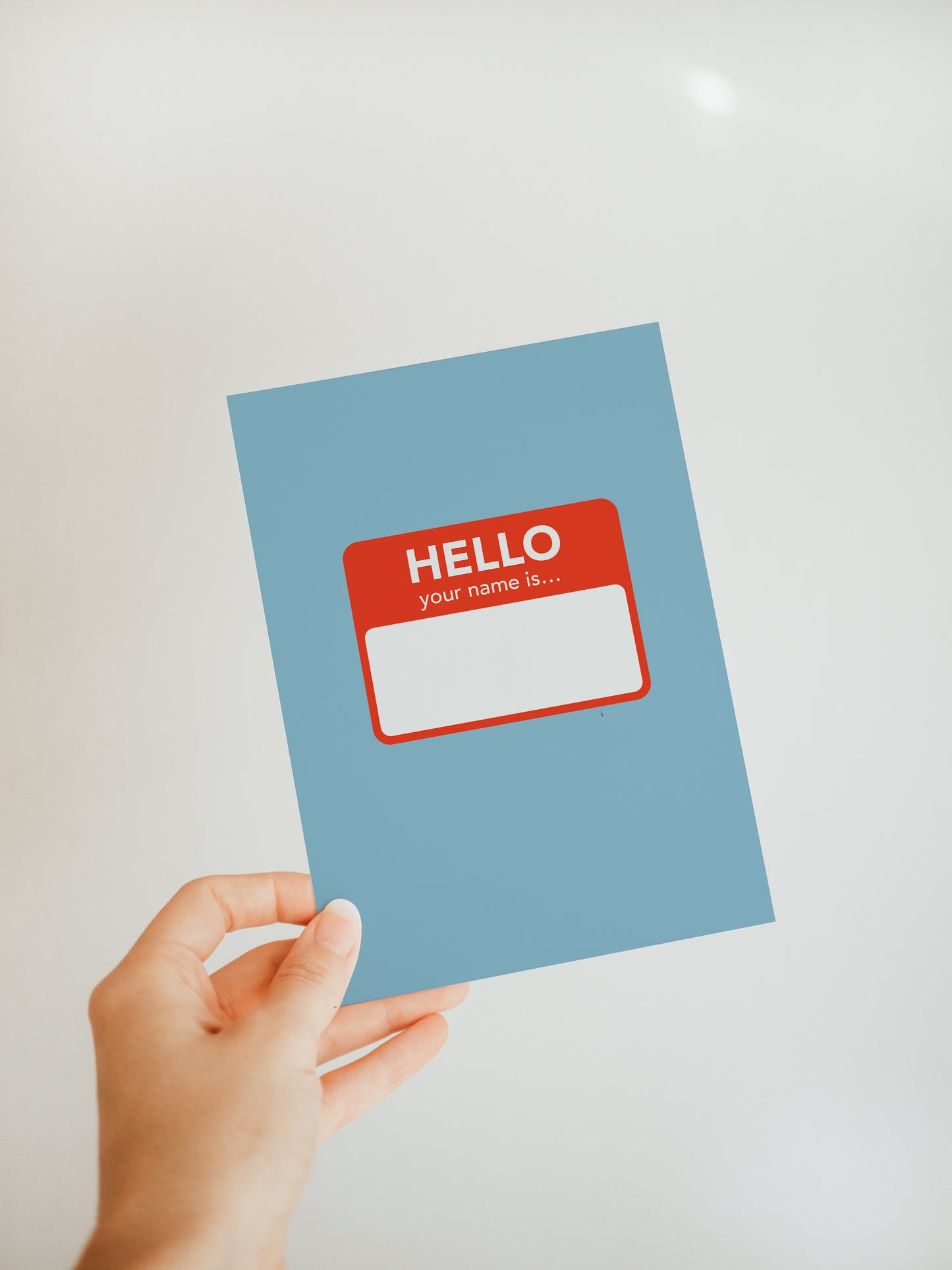 hand holding a blue greeting card with a design resembling a red and white name tag sticker in the center that says "hello, your name is"