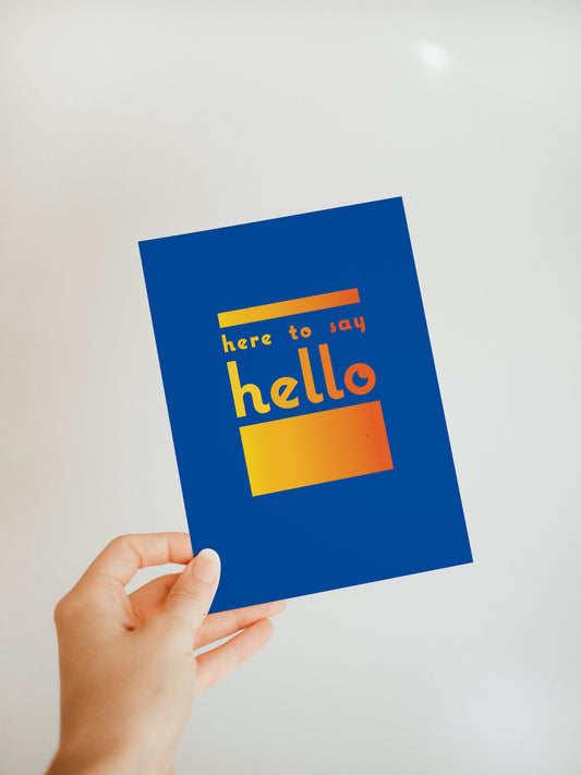 hand holding a blue greeting card that says "here to say hello" in a gradient red and orange font with a block of the same gradient colors above and below the words