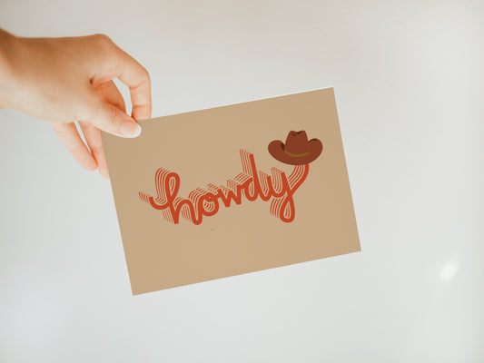 hand holding a tan greeting card with dark red letters that spell "howdy" with a cowboy hat sitting at the end of the word