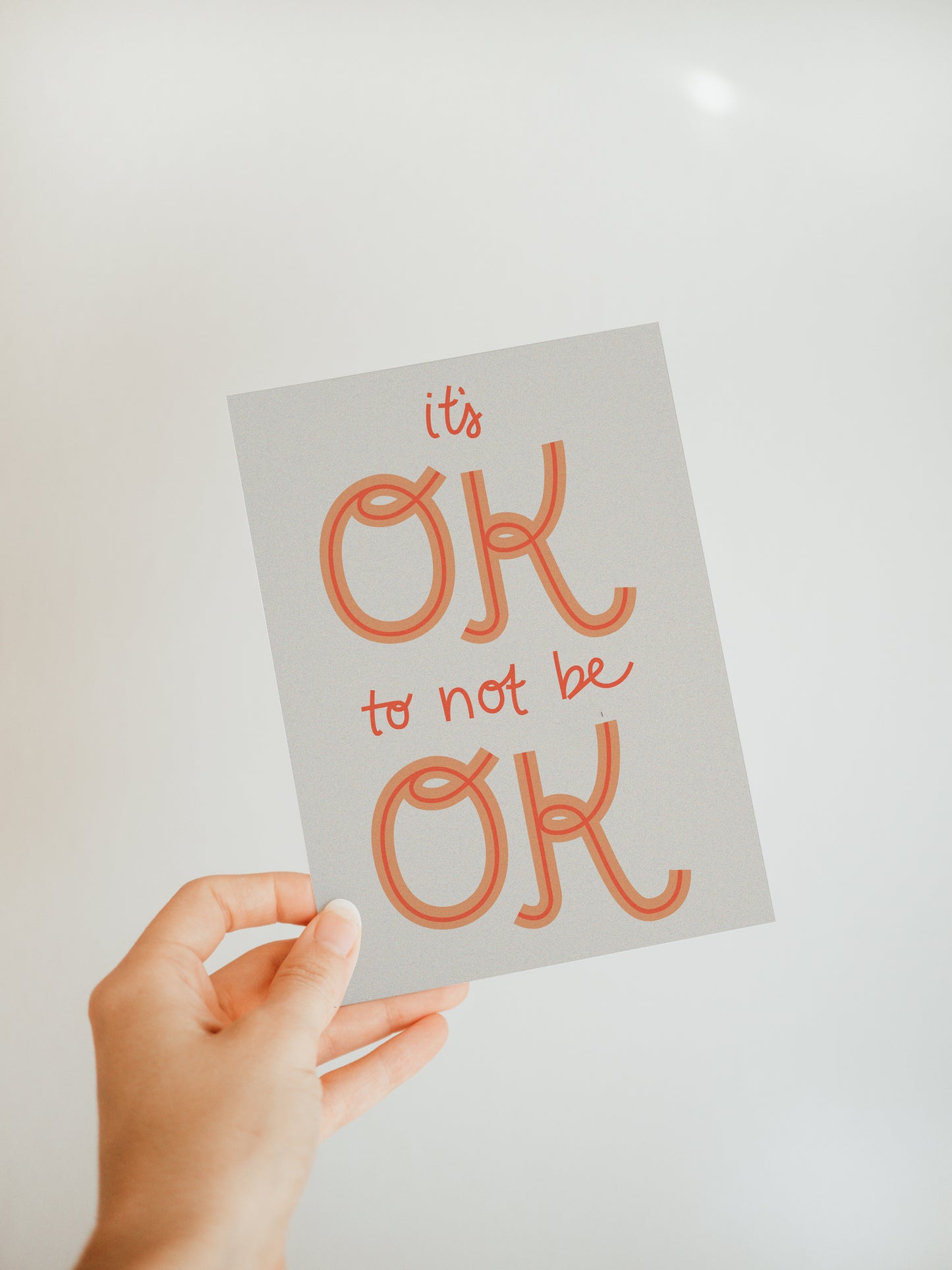 hand holding an off-white greeting card that says "it's ok to not be ok" in dark orange font