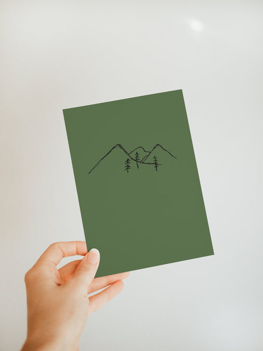 hand holding a forest green greeting card with a simple mountain range sketched design and three small pine trees in front of the mountains