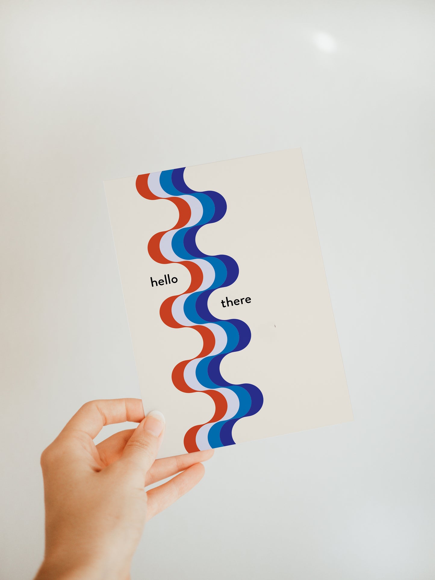 hand holding a white greeting card with a red and blue ribbon like design vertically down the front and small words saying  "hello there" on either side of the ribbon