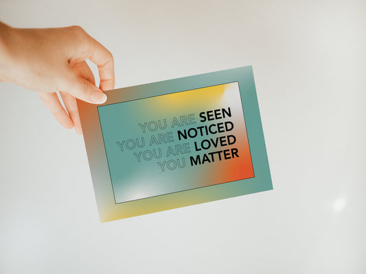 hand holding a multicolored greeting card that says "you are seen, you are noticed, you are loved, you matter."