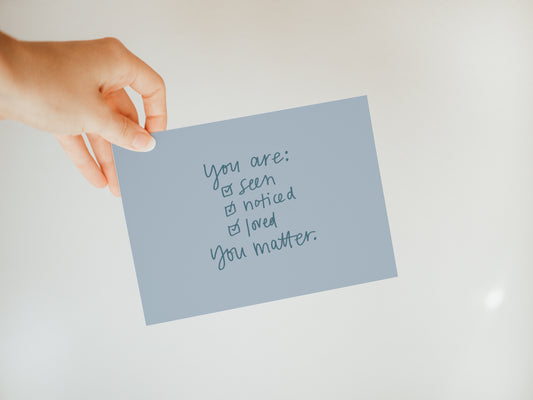 hand holding a dusky blue greeting card that has a checklist and says "you are: seen, noticed, loved, you matter."
