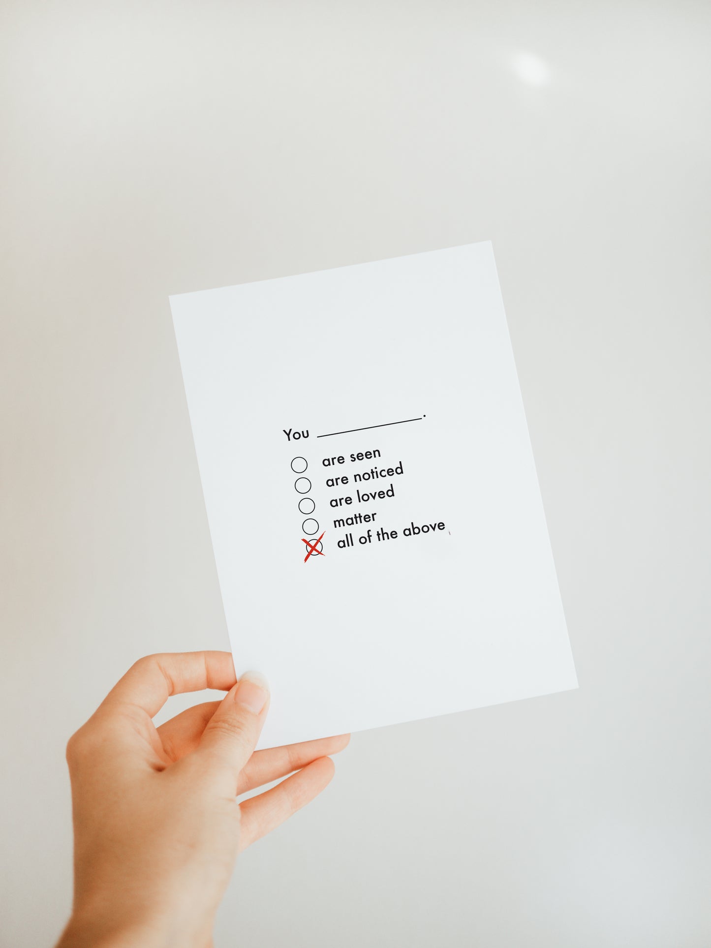 hand holding a white greeting card with the words "you" followed by check boxes with the options "are seen, are noticed, are loved, matter, all of the above" the last option of "all of the above" is checked with a red mark