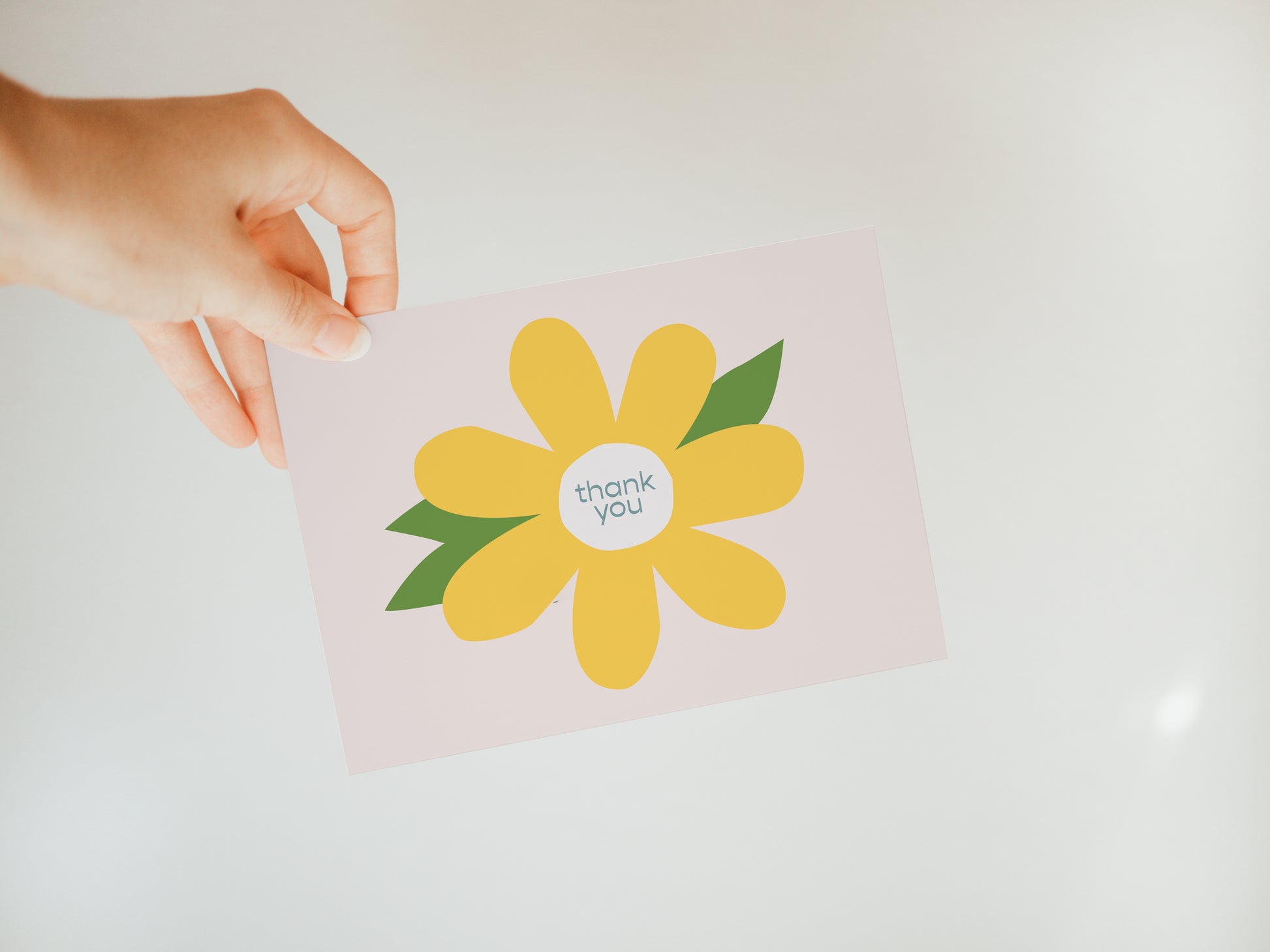 hand holding a light pink greeting card with a large yellow flower and the words "thank you" in the flower's center