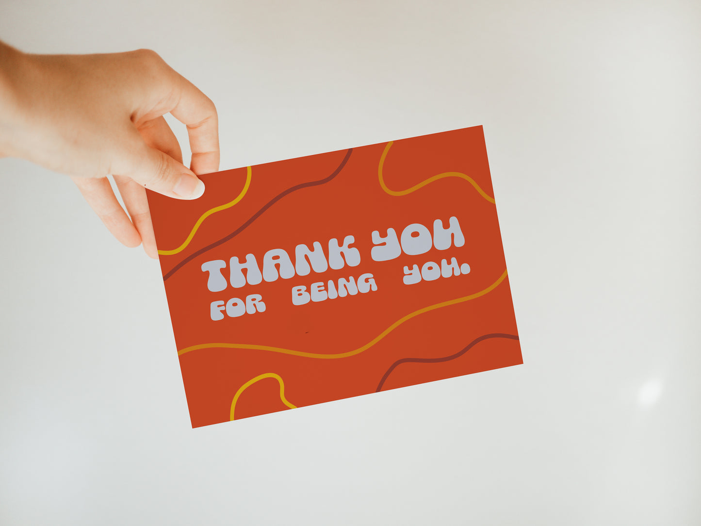 hand holding a red greeting card that says "thank you for being you" in white bubble letters with a simple wavy line design in the background in muted yellows and dark reds
