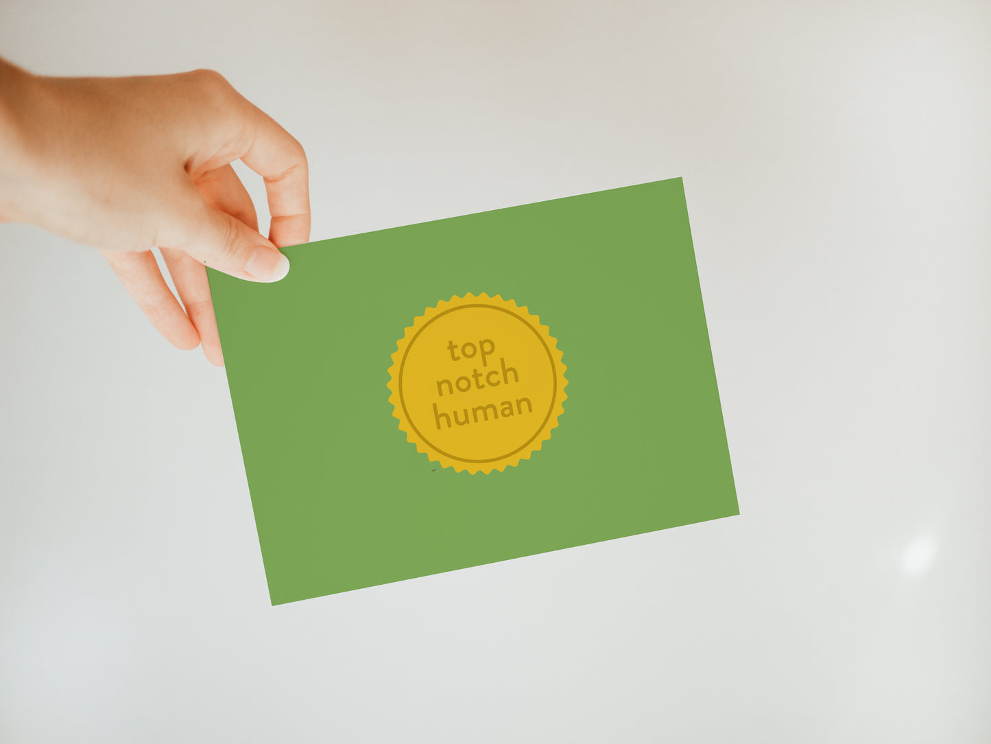 hand holding a green greeting card with a yellow design in the middle that looks like a medal and says "top notch human"
