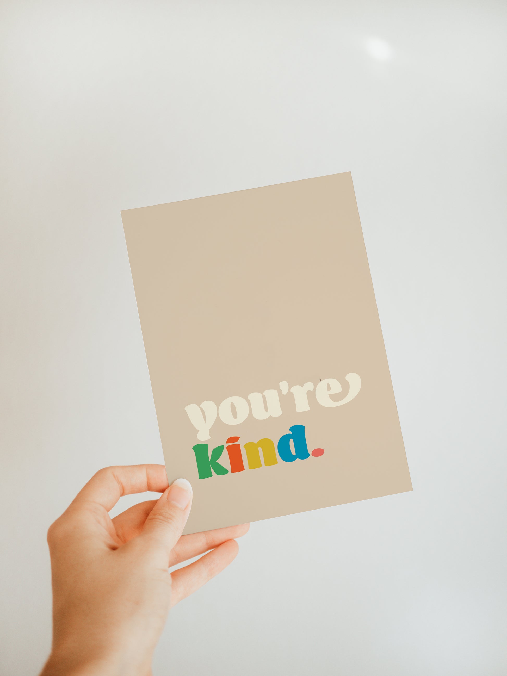 hand holding a dark cream greeting card with the words "you're kind" on the bottom in white and multicolored font