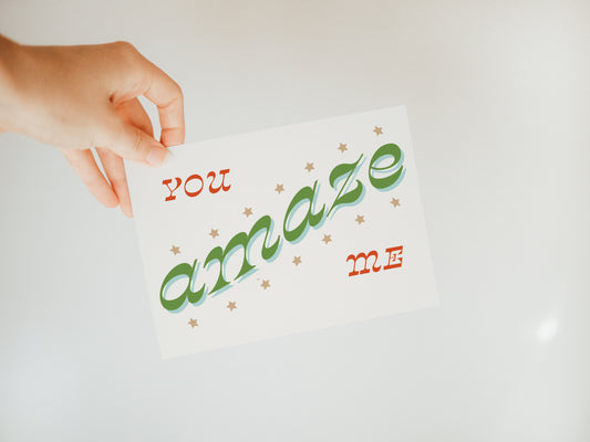hand holding an off-white greeting card with the words "you amaze me" in red and green fonts with stars around the word "amaze"