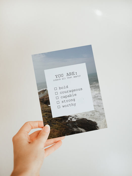 hand holding a greeting card with an ocean scene in the background and a white square in the foreground that says "you are: (check all that apply) bold, courageous, capable, strong, worthy."