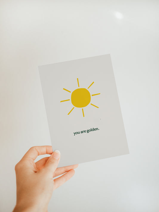 hand holding an off-white greeting card with a golden yellow sun and the words "you are golden" in black